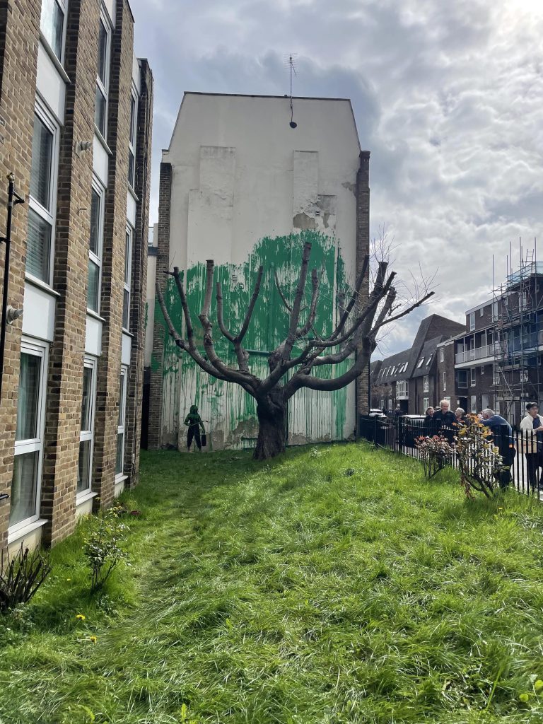 Banksy green tree mural beside a pruned cherry tree on Hornsey Road