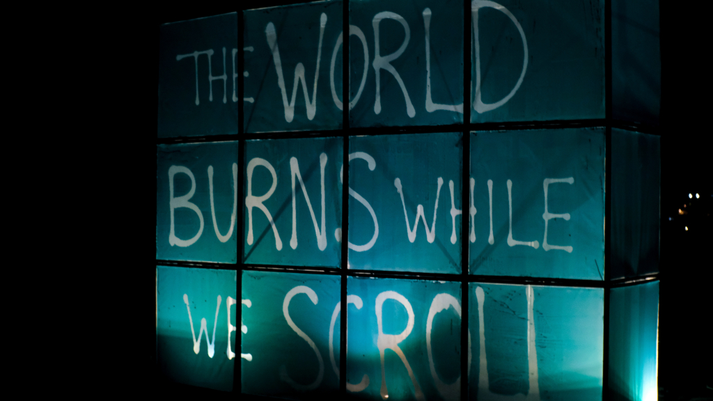 Illuminated words “THE WORLD BURNS WHILE WE SCROLL” across Ricardo Romero’s painting-screen installation.