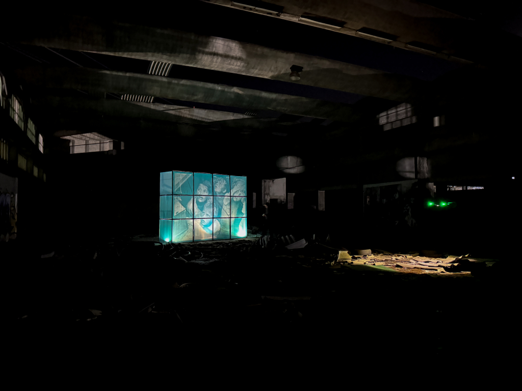 Wide view of Ricardo Romero’s illuminated wartime installation surrounded by the dark architecture of an industrial building.